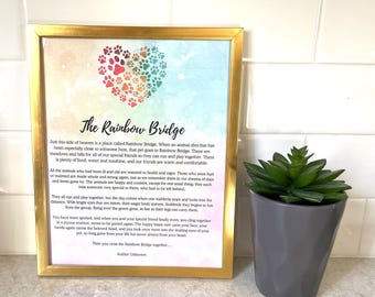 Puede incluir: Una impresión enmarcada con borde dorado presenta el texto "The Rainbow Bridge" y un poema sobre mascotas. Un corazón hecho de huellas de patas coloridas está en la parte superior. Una pequeña planta suculenta verde en una maceta gris está al lado.
