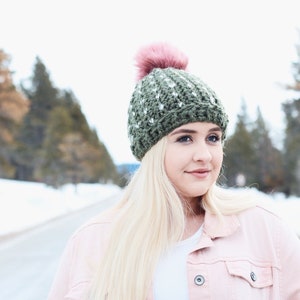 May include: A woman wearing a green and white crocheted beanie with a pink pom pom. She is wearing a pink jacket and has long blonde hair.