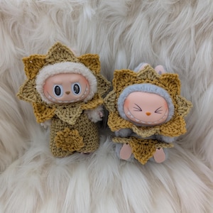 May include: Two small dolls, each with a round face and large eyes, are dressed in crocheted sunflower costumes. The costumes are yellow and tan, with a white fluffy trim around the face. The dolls are on a white, fluffy surface.