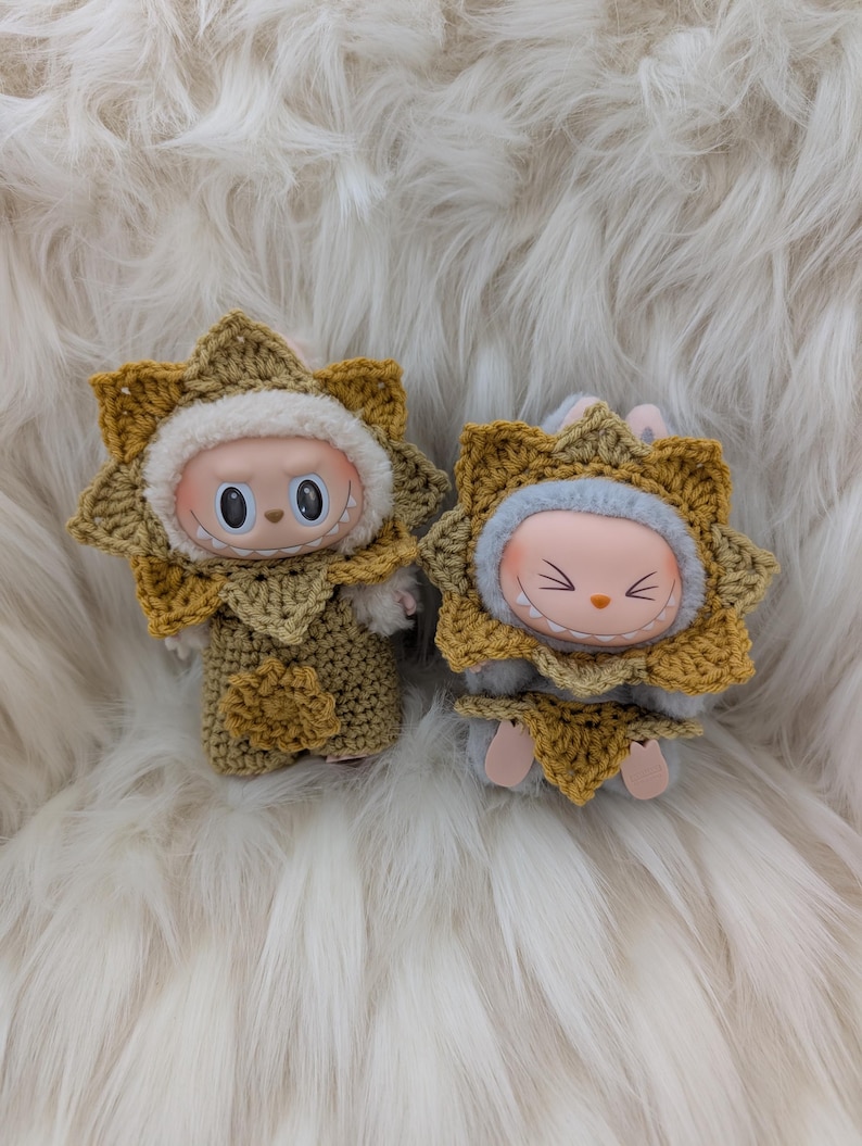 May include: Two small dolls, each with a round face and large eyes, are dressed in crocheted sunflower costumes. The costumes are yellow and tan, with a white fluffy trim around the face. The dolls are on a white, fluffy surface.