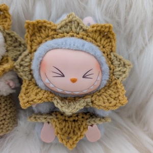 May include: A charming figurine with a smiling face, dressed in a handmade crochet outfit in shades of yellow and tan. The outfit resembles a lion's mane, with a light blue trim around the face. The figurine has small pink feet.
