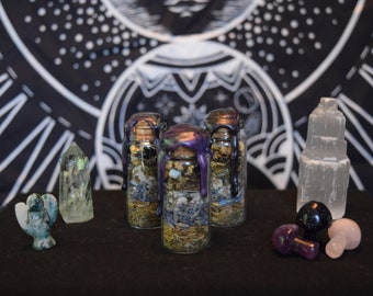 May include: Three small glass bottles filled with herbs and crystals, sealed with a purple wax. The bottles are arranged in a triangle shape on a black surface. There are several other crystals and a small green figurine on the surface.