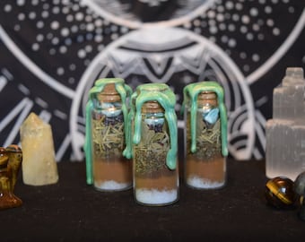 May include: Three small glass bottles filled with herbs, crystals, and a brown powder. The bottles are sealed with cork stoppers and topped with a green wax seal. The bottles are arranged in a row on a black surface.