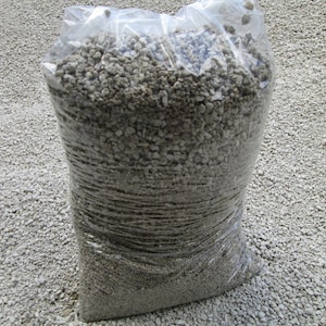 May include: A clear plastic bag filled with small, light gray, irregularly shaped rocks. The bag is sitting on a surface of similar rocks.