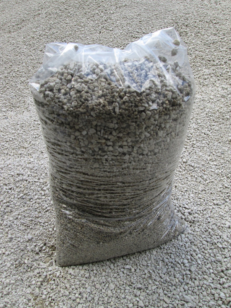 May include: A clear plastic bag filled with small, light gray, irregularly shaped rocks. The bag is sitting on a surface of similar rocks.