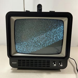 May include: A black RCA portable television with a silver screen bezel and a telescoping antenna. The screen displays static noise. The front panel features a speaker grille and two silver control knobs labeled BRIGHT and ON.
