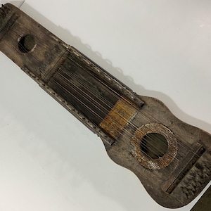 Pode incluir: Um instrumento de cordas de madeira desgastada, com corpo retangular e seção inferior arredondada. Apresenta um orifício sonoro circular e inúmeras cordas, exibindo uma estética rústica e antiga. A madeira tem uma aparência escura e envelhecida.