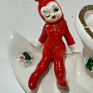 May include: A vintage ceramic figurine of a winking elf in a red jumpsuit and hat, seated on a white crescent-shaped base. The base is decorated with small flowers. The figurine has white gloves and a painted face.