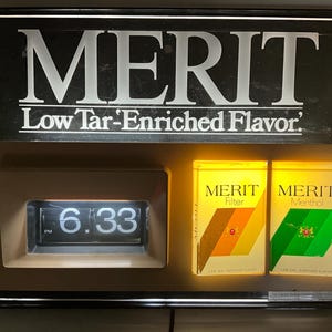 May include: A vintage Merit cigarette display with a digital clock displaying 6:33 PM. The sign reads "MERIT Low Tar-Enriched Flavor." Two illuminated cigarette packs are visible.