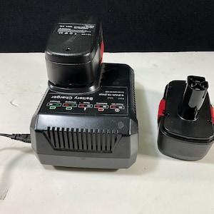 以下が含まれることがあります： 黒いバッテリー充電器と、取り付けられた黒いバッテリー、および赤いアクセントが付いた別の黒いバッテリー。 充電器にはインジケーターライトと「Battery Charger」の文字があります。 充電器はコンセントに接続されています。