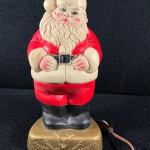 Peut inclure: Une figurine de Père Noël en plastique vintage avec un costume rouge, une barbe blanche et des bottes noires. La figurine tient un sac et possède une fonction d'éclairage. La base de la figurine est dorée et comporte le texte "Joyeux Noël" et un renne.