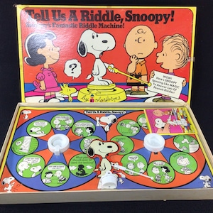 May include: A vintage board game featuring Snoopy from the Peanuts comic strip. The game is called "Tell Us A Riddle, Snoopy!" and features a spinning wheel with different riddles. The game is in a box with a colorful illustration of Snoopy and other Peanuts characters.