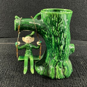 May include: A green ceramic planter shaped like a tree trunk with a small elf figurine sitting on a swing attached to the tree. The elf is wearing a green outfit and has a white face.