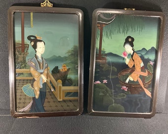May include: Two framed paintings depicting women in traditional Chinese clothing. The painting on the left shows a woman standing on a balcony with a fan in her hand. The painting on the right shows a woman sitting in a boat with a lotus flower in her hand.