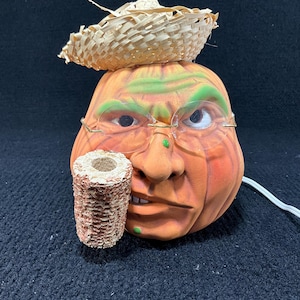 Könnte beinhalten: Halloween-Dekoration mit einem Kürbiskopf, Strohhut und Brille. Der orangefarbene Kürbis hat grüne Akzente und ein detailliertes Gesicht. Eine Maiskolbenpfeife wird im Mund gehalten. Ein weißes Elektrokabel erstreckt sich von der Rückseite.