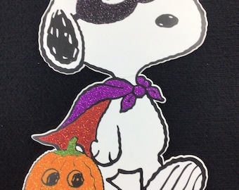 Op de afbeelding: Een witte cartoon hond met een paarse cape en een zwart oogmasker houdt een oranje pompoen vast met een pompoengezicht. De hond staat op een witte achtergrond. ©2011 Peanuts Worldwide LLC