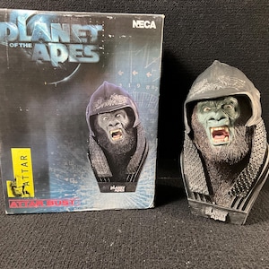 May include: A black and gray bust of the character Attar from the movie Planet of the Apes. The bust is on a black base with the words "Planet of the Apes" printed on it. The bust is in a box with the words "Planet of the Apes" and "Attar Bust" printed on it.