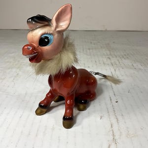 Könnte beinhalten: Vintage Keramik-Eselfigur mit rotem Körper, beigem Fellkragen und kleinem weißem Schwanz. Der Esel hat große blaue Augen, rosa Ohren und eine schwarz-beige Mähne. Die Figur ist in einer stehenden Position dargestellt.