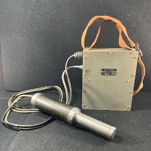 May include: An antique, olive-drab metal device with a cylindrical microphone and a rectangular box with a shoulder strap. The microphone is dark gray with a silver tip. The box has a metal plate with text. Cables connect the microphone to the box.