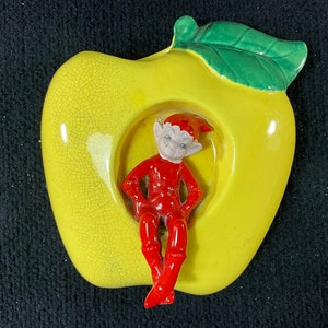 Pode incluir: Uma maçã de cerâmica amarela com uma folha verde e uma pequena figura de elfo vermelho sentada dentro. A maçã tem um esmalte craquelado.