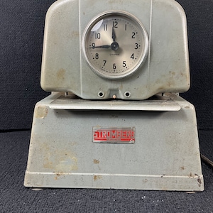Könnte beinhalten: Eine alte graue Metall-Zeituhr mit weißem Zifferblatt und schwarzen Zeigern. Das Zifferblatt hat schwarze Zahlen und den Text "Stromberg" in Schwarz. Die Uhr ist auf einem grauen Metallsockel mit dem Text "Stromberg Model No. 10" in Rot montiert.