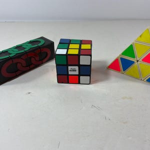 Può includere: Una collezione di tre puzzle che include un blocco rettangolare nero con grafiche di maglie di catena verdi e rosse, un cubo di Rubik standard con facce quadrate multicolori e un puzzle a piramide triangolare giallo, verde, blu e rosa.