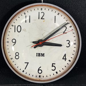 May include: Vintage round wall clock with a white face, black numerals, and black hands. The clock has a metal frame and a hanging loop at the top. The clock face has the IBM logo and the words "MADE IN U.S.A."