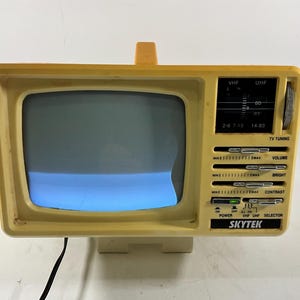 May include: A vintage yellow Skytek portable television with a small screen. The TV has a handle, tuning dials, and control knobs for volume, brightness, and contrast. The screen displays a blue hue. The text "SKYTEK" is visible.