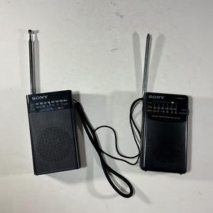 May include: Two black Sony portable radios with extending aerials. The radios have a speaker grille, tuning dials, and a wrist strap. The radios are on a white surface.