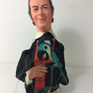 May include: A vintage hand puppet of a man with a black suit and a plaid shirt. He is holding a top hat and a blue parrot is perched on the hat.