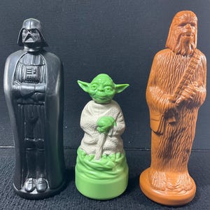 Puede incluir: Tres figuras de personajes de Star Wars: Darth Vader en negro, Yoda en verde y blanco y Chewbacca en marrón. Las figuras están de pie y parecen ser de plástico.