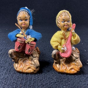 May include: Two vintage ceramic figurines. One figure wears a blue outfit and plays red drums. The other figure wears a yellow outfit and plays a pink stringed instrument. Both figures sit on a brown base.