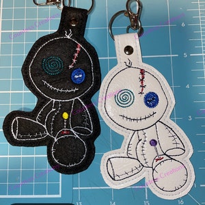 May include: Two felt voodoo doll keychains. One is black with yellow and blue details. The other is white with blue and red details. Both have a stitched-on smile and a single button eye.  The keychains are on a blue cutting mat with a ruler.