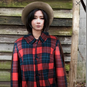 Könnte beinhalten: Eine Frau trägt eine rot-schwarze karierte Jacke mit einem braunen Filzhut. Sie trägt einen schwarzen Rock und schwarze Strumpfhosen.