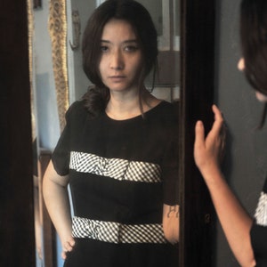 Puede incluir: Una mujer con un vestido de cuadros vichy blanco y negro se para frente a un espejo. El vestido tiene un corpiño negro y un ribete de cuadros vichy blanco alrededor del escote, la cintura y el dobladillo.