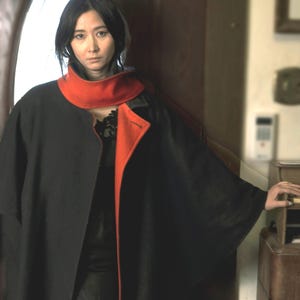 May include: A black cape featuring a vibrant orange inner lining and a high, structured collar. The garment is worn over a black top, showcasing a contrasting color scheme with a wide, open front design and loose, flowing sleeves.