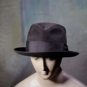 May include: A dark brown fedora hat with a black band. The hat has a classic, slightly rounded crown and a wide brim. The hat is displayed on a mannequin.