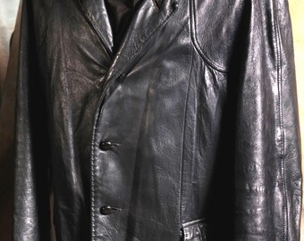May include: A black leather jacket featuring a notched collar, long sleeves, and a front button closure with three visible black buttons. The jacket has a textured surface, structured shoulders, and a small flap pocket detail on the lower left side.