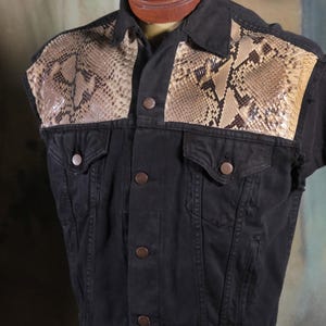 May include: A black sleeveless denim vest featuring tan and brown snakeskin-patterned panels across the upper chest and shoulders. The vest includes a classic collar, a front button closure with metallic buttons, and two chest pockets.