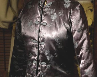 May include: A dark purple satin jacket featuring a mandarin collar, long sleeves, and a repeating floral pattern in a lighter shade. The garment closes with five fabric-covered toggle buttons down the front and includes two horizontal pockets.