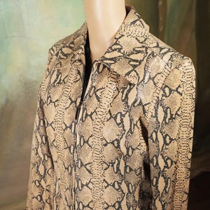 Puede incluir: Una chaqueta de manga larga con un estampado de piel de serpiente en tonos beige y negro. La prenda tiene un cuello puntiagudo, un cierre de cremallera frontal completo y un corte estructurado, exhibida sobre un maniquí para mostrar el cuello.