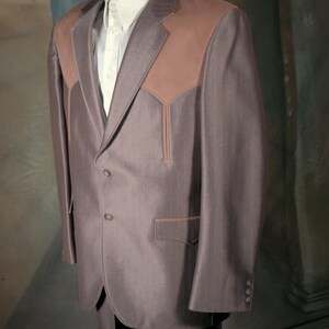 May include: A brown suit jacket featuring a textured pinstripe pattern, solid brown yokes on the shoulders, a two-button front closure, and two lower pockets with brown trim. The jacket is worn over a light-coloured button-down shirt with a collar.