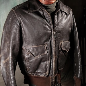 May include: A dark brown leather flight jacket featuring a front zip closure, a pointed collar, and two front flap pockets with button fastenings. The jacket includes ribbed brown knit cuffs and a matching ribbed hem, showing a worn texture.