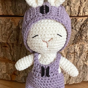 May include: A handmade crochet bunny doll with a white body and lavender overalls and hat. The bunny has long white ears and a smiling face. The overalls have black accents. The doll is standing on a wooden surface.
