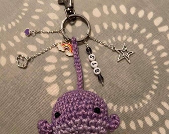 May include: A handmade purple crochet whale keychain with a silver clasp. The keychain features charms including a star, a rainbow, a heart, and a paw print. The letters "BTS" are spelled out on beads.