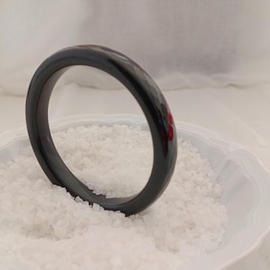 Könnte beinhalten: Ein glänzendes, schwarzes Armreif-Armband. Das kreisförmige Armband besteht aus einem glatten, festen Material. Es liegt auf einem Bett aus weißem, körnigem Material in einer weißen Schale mit gewelltem Rand. Das Armband hat einen Durchmesser von ca. 7 cm.