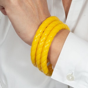 Op de afbeelding: Vier gele plastic armbanden gestapeld op een pols. De armbanden zijn dik en hebben een geribbelde textuur.