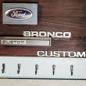 Puede incluir: Un llavero de madera con el logotipo de Ford, la palabra "BRONCO" y la palabra "CUSTOM" en letras plateadas. El llavero tiene seis ganchos negros y un esquema de color rústico, marrón y blanco.