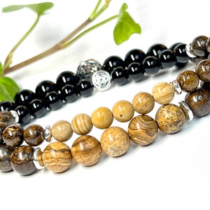 May include: Two beaded bracelets, one with black beads and the other with brown and beige beads. The black beaded bracelet has a silver charm in the shape of a circle with a design inside.