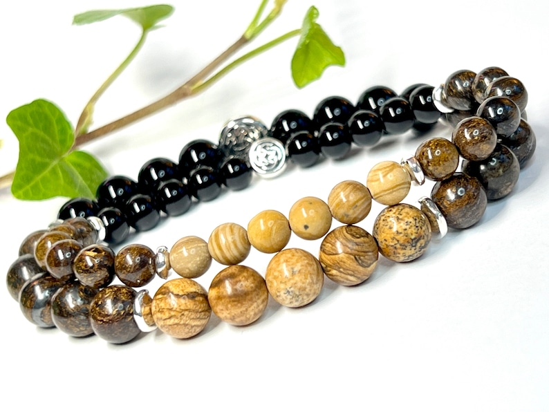 May include: Two beaded bracelets, one with black beads and the other with brown and beige beads. The black beaded bracelet has a silver charm in the shape of a circle with a design inside.